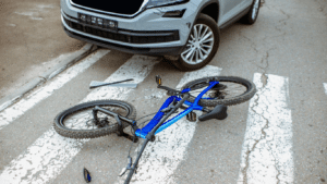 A blue bicycle lies on a crosswalk in front of a silver car with a damaged bumper, suggesting a recent collision.