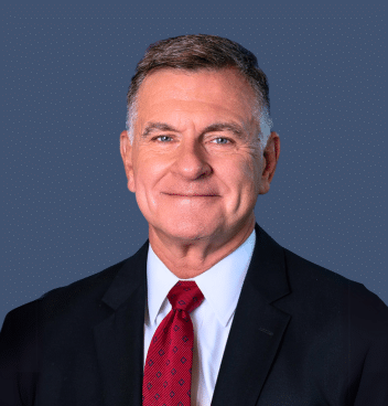 A middle-aged man in a dark suit, white shirt, and red tie, smiling, posed against a plain blue background.