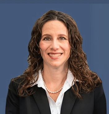 A woman with long curly brown hair, wearing a black blazer, white blouse, and pearl necklace, smiles in front of a solid blue background.