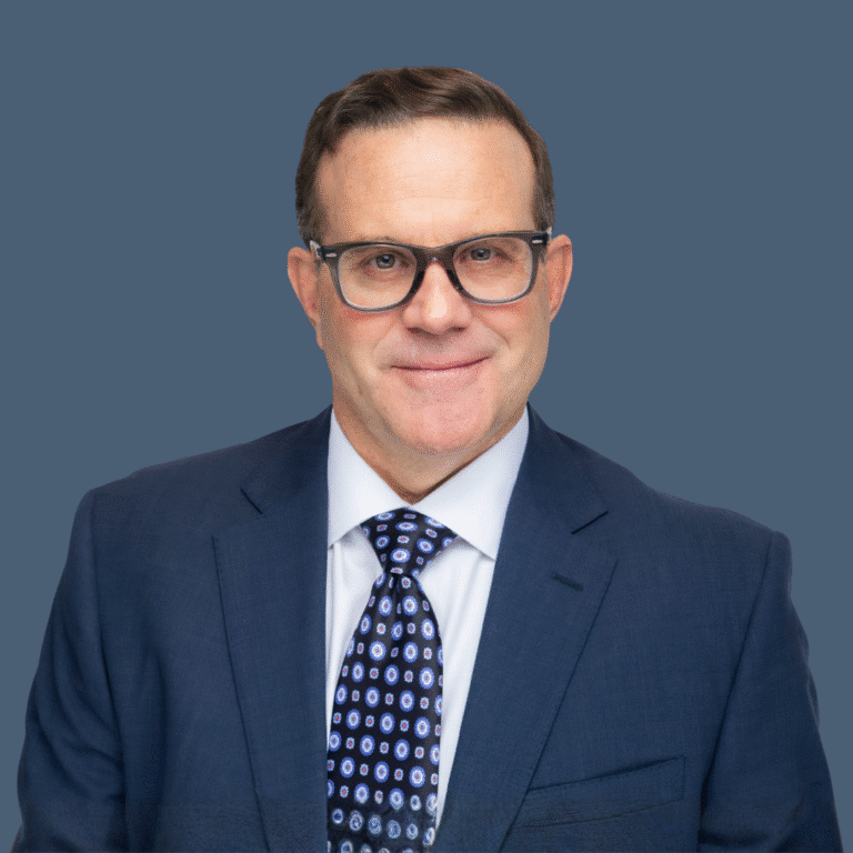 A man wearing glasses, a blue suit, white shirt, and patterned tie poses against a plain blue background.
