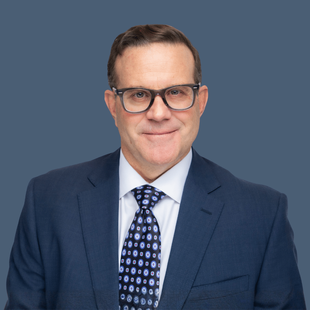 A man wearing glasses, a blue suit, white shirt, and patterned tie poses against a plain blue background.