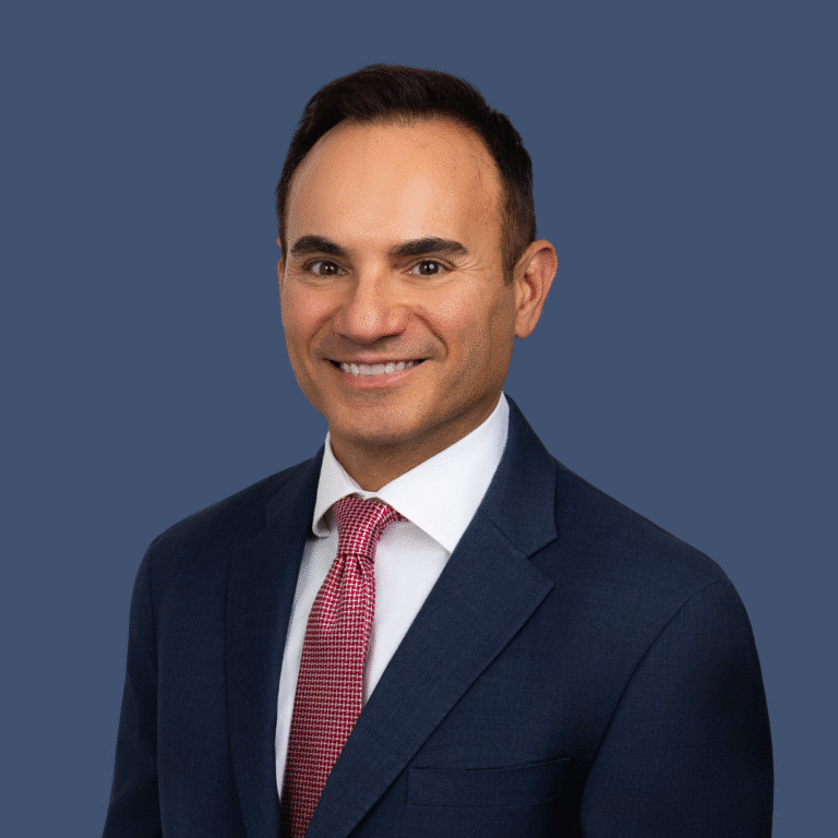 A man in a navy suit, white shirt, and red patterned tie smiles at the camera against a plain blue background.