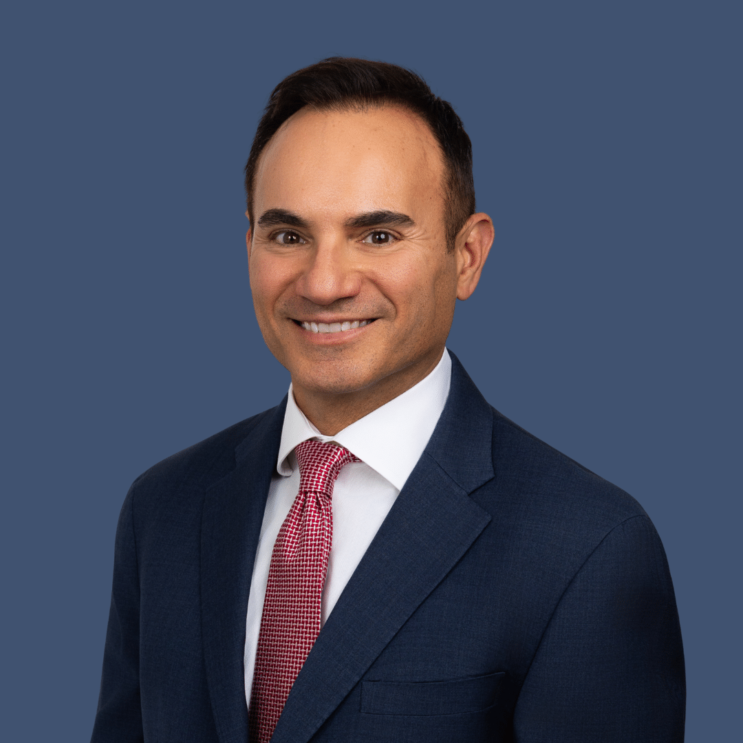 A man in a navy suit, white shirt, and red patterned tie smiles at the camera against a plain blue background.