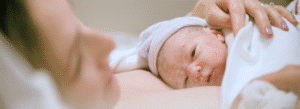 A newborn baby wearing a striped hat lies on a woman’s chest, partially covered by a white blanket. The woman’s hand rests gently on the baby.