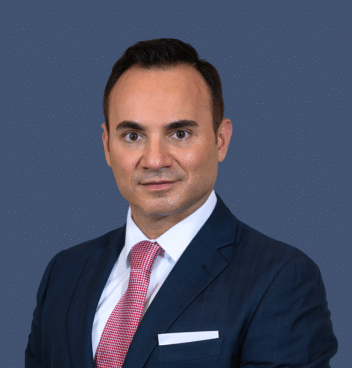 A man in a navy suit, white shirt, and pink tie poses for a formal portrait against a plain blue-gray background.