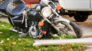 A black motorcycle lies on its side on grass near a bent pole, with a car and a truck parked in the background.