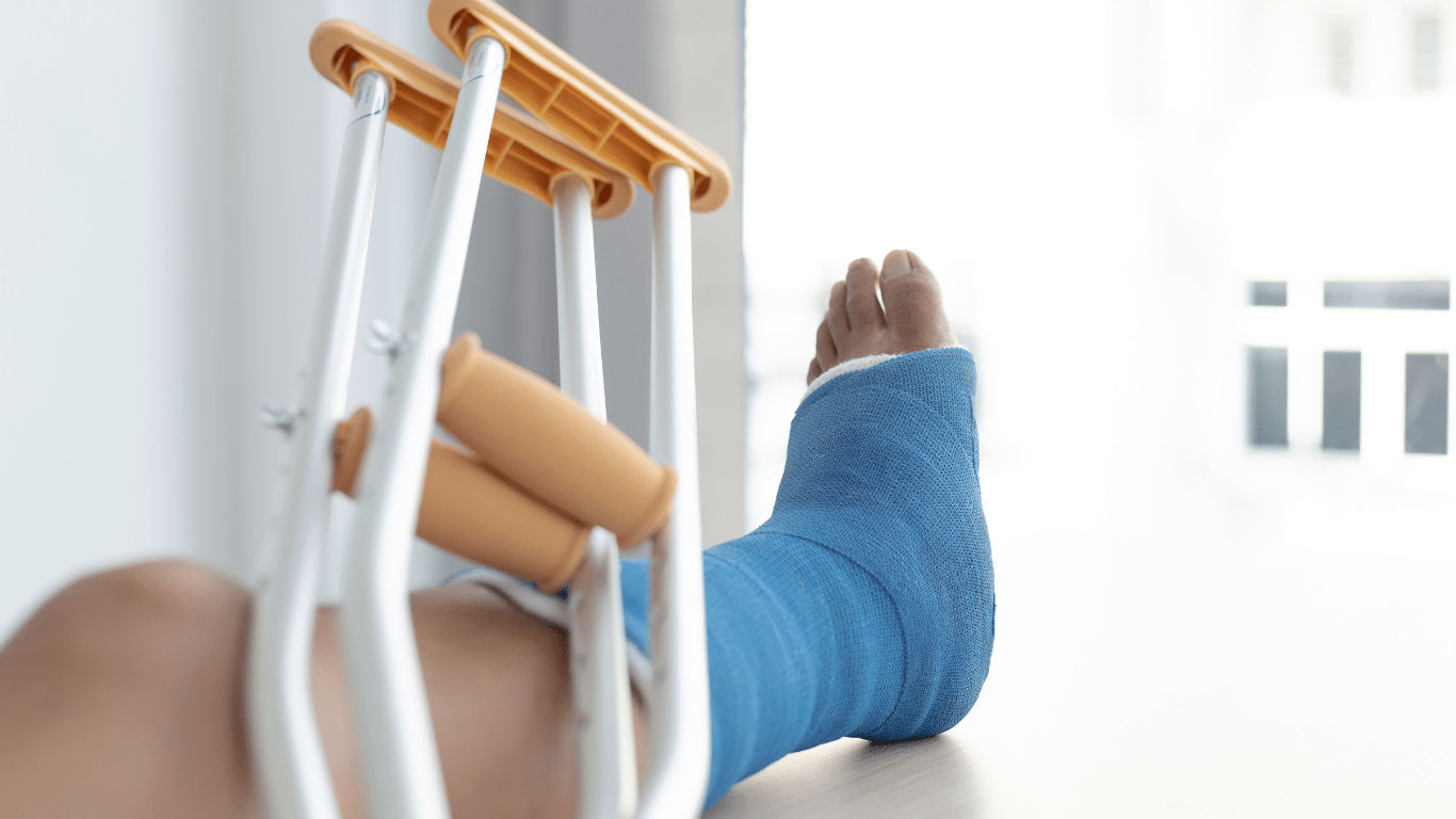 A person’s leg in a blue cast rests on a surface next to a pair of crutches, with natural light coming through a window in the background.