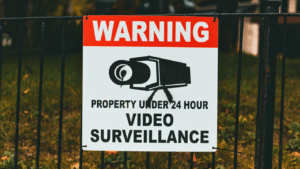 A warning sign on a fence reads “Property under 24 hour video surveillance” with an image of a security camera on it.