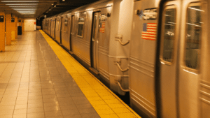 A silver subway train with an American flag moves through an underground station with yellow safety tiles and empty platforms.