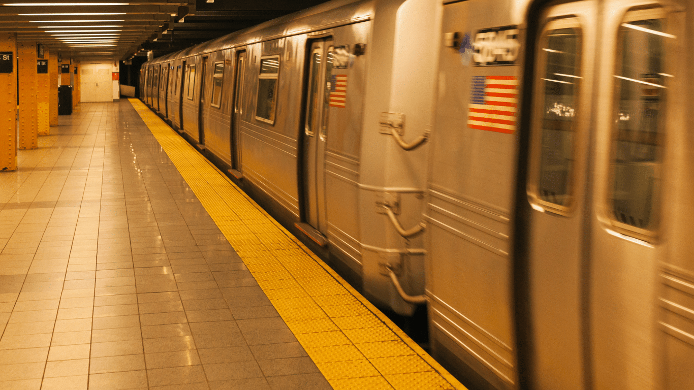 A silver subway train with an American flag moves through an underground station with yellow safety tiles and empty platforms.