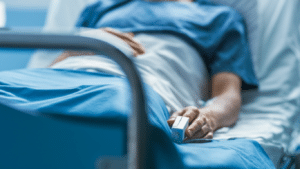 Patient lies in a hospital bed, wearing a gown with a pulse oximeter on their finger, in a clinical setting.