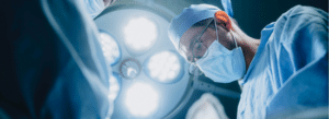 A surgeon wearing a mask and surgical cap performs an operation under bright operating room lights.