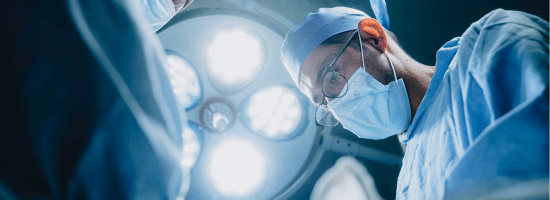 A surgeon wearing a mask and surgical cap performs an operation under bright operating room lights.
