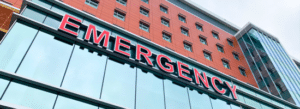 A modern hospital building with large red "EMERGENCY" sign above the glass entrance.