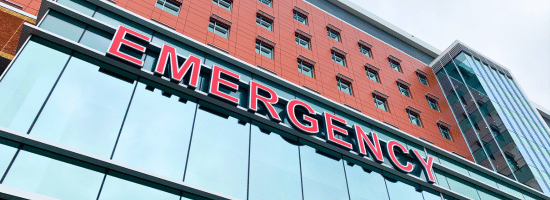 A modern hospital building with large red "EMERGENCY" sign above the glass entrance.