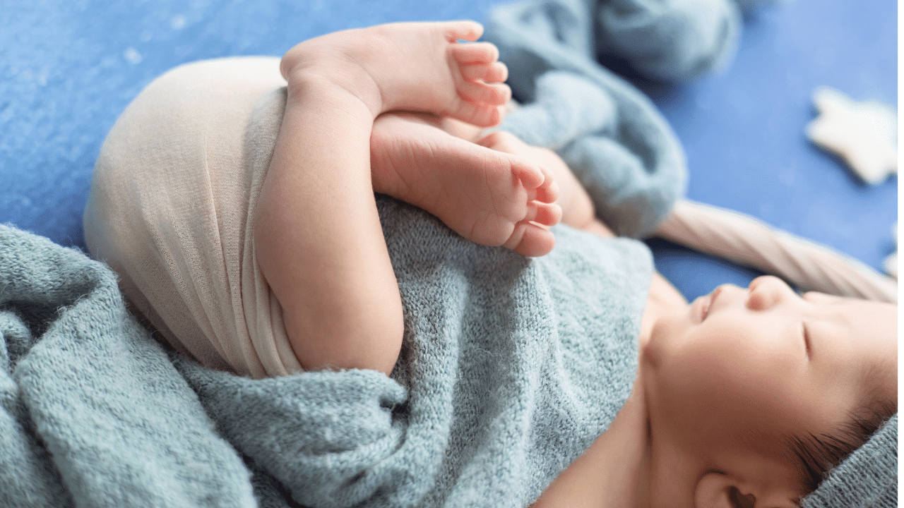 A baby lying on its back, wrapped in a soft blue blanket, with legs bent and feet touching, resting on a blue surface.