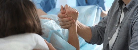 A person in a hospital bed holds hands with a companion, with medical staff in blue scrubs assisting in the background.