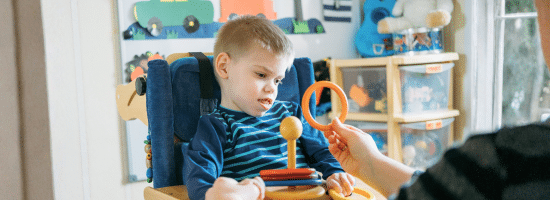 A young boy in a specialized chair looks at a person holding a plastic ring in a playroom with toys and educational materials in the background.