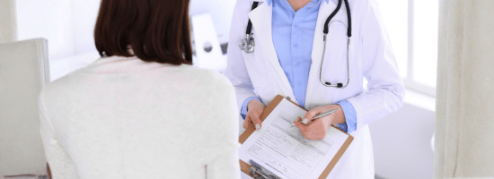 A doctor in a white coat holds a clipboard and talks to a patient seated across from them.