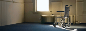 A wheelchair sits in an empty, dimly lit room near a window with sunlight streaming in and a radiator beneath the window.