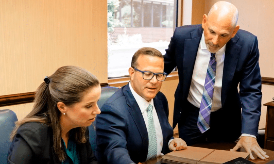 Three individuals in professional attire are seated at a table, discussing documents. An office setting is visible in the background.