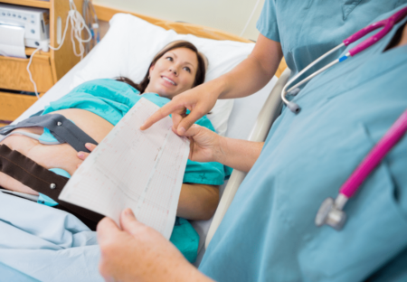 A pregnant woman lies in a hospital bed with fetal monitoring belts on her abdomen. A healthcare professional shows her a printout, explaining the results.