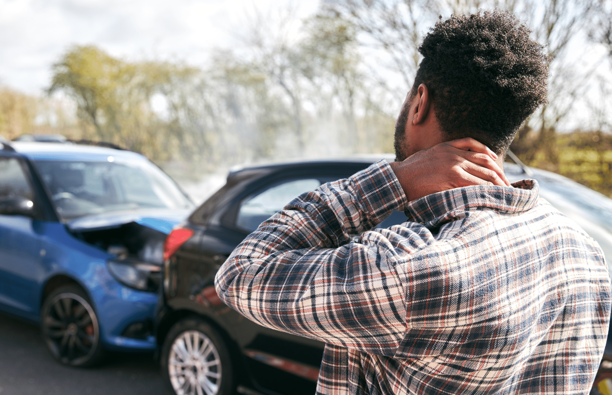 A man holds his neck while standing near two cars involved in a rear-end collision on a road.