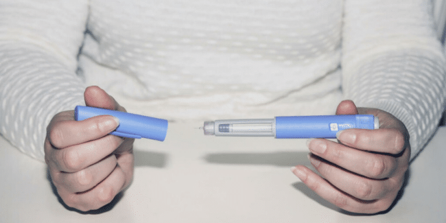 Person holding an insulin pen with the cap removed, wearing a white textured long-sleeve shirt, sitting at a light-colored surface.