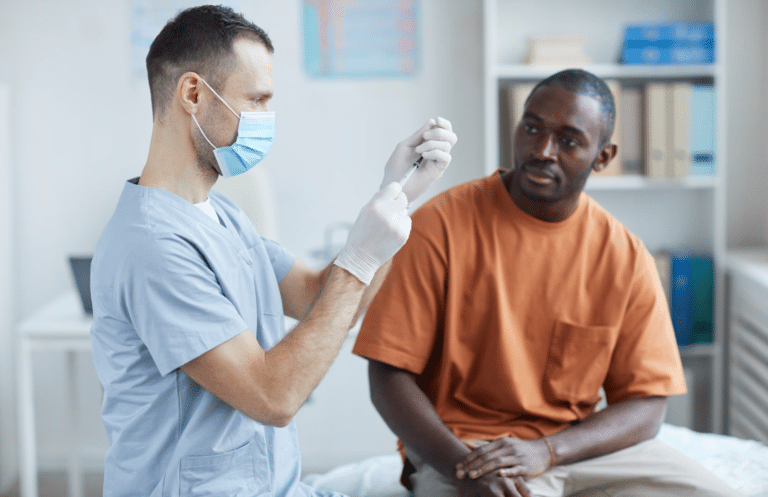 A healthcare worker wearing scrubs and a mask prepares a syringe while a patient in an orange shirt sits nearby and watches in a medical office.