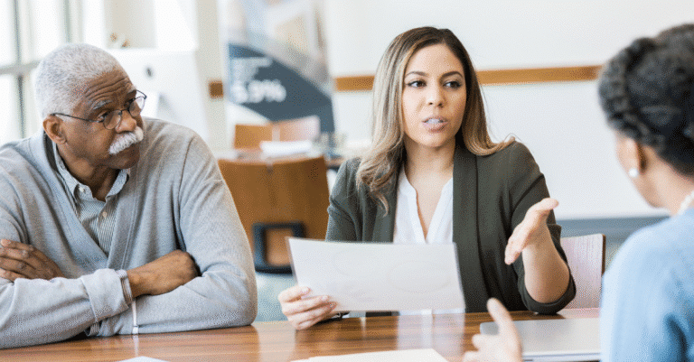 Three people sit at a table having a discussion; a woman in the center holds papers and gestures while talking, while the others listen.