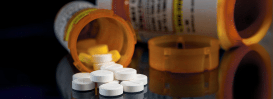 Several white tablets are scattered on a reflective surface beside an open prescription pill bottle, with another bottle lying on its side in the background.