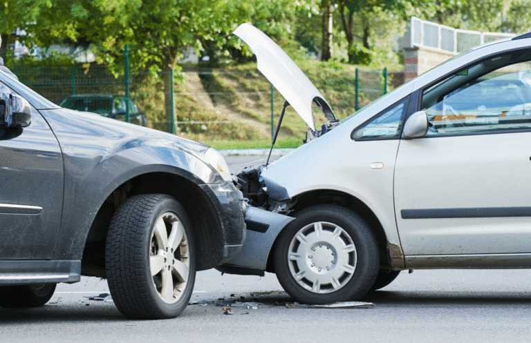 Two cars involved in a front-end collision on a road, with the hood of one vehicle open and visible damage to both cars. Trees and a fence are in the background.