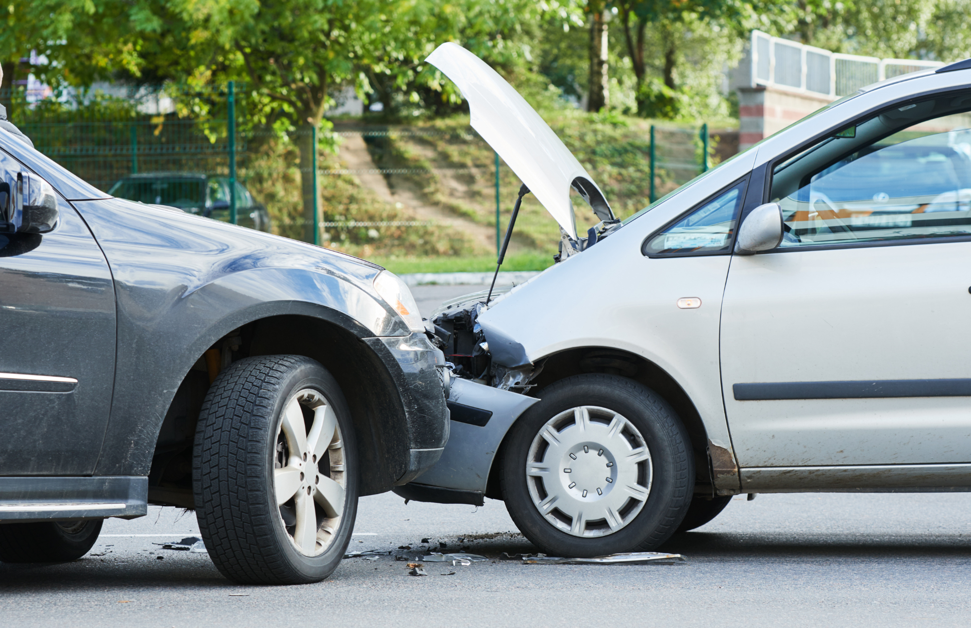 Two cars involved in a front-end collision on a road, with the hood of one vehicle open and visible damage to both cars. Trees and a fence are in the background.