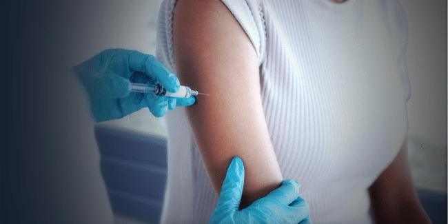 A healthcare worker wearing blue gloves administers an injection to a person’s upper arm.