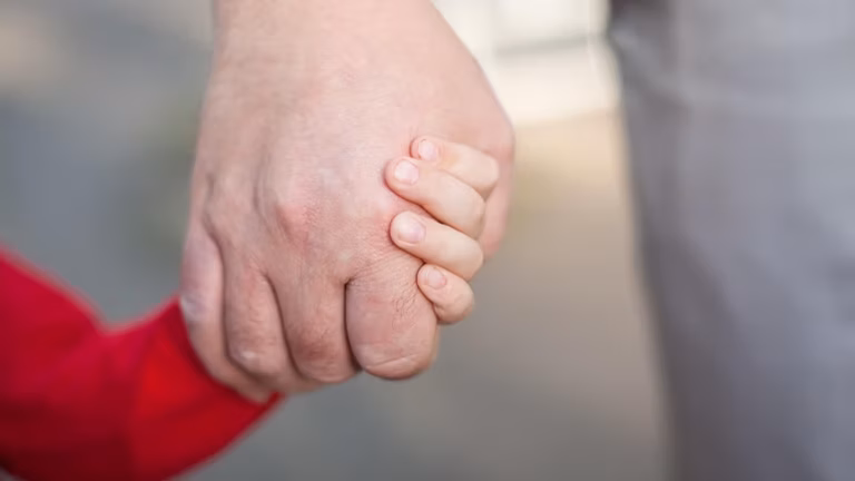 Close-up of a child’s hand holding an adult’s hand, with both facing forward. The child is wearing a red sleeve. The background is blurred.