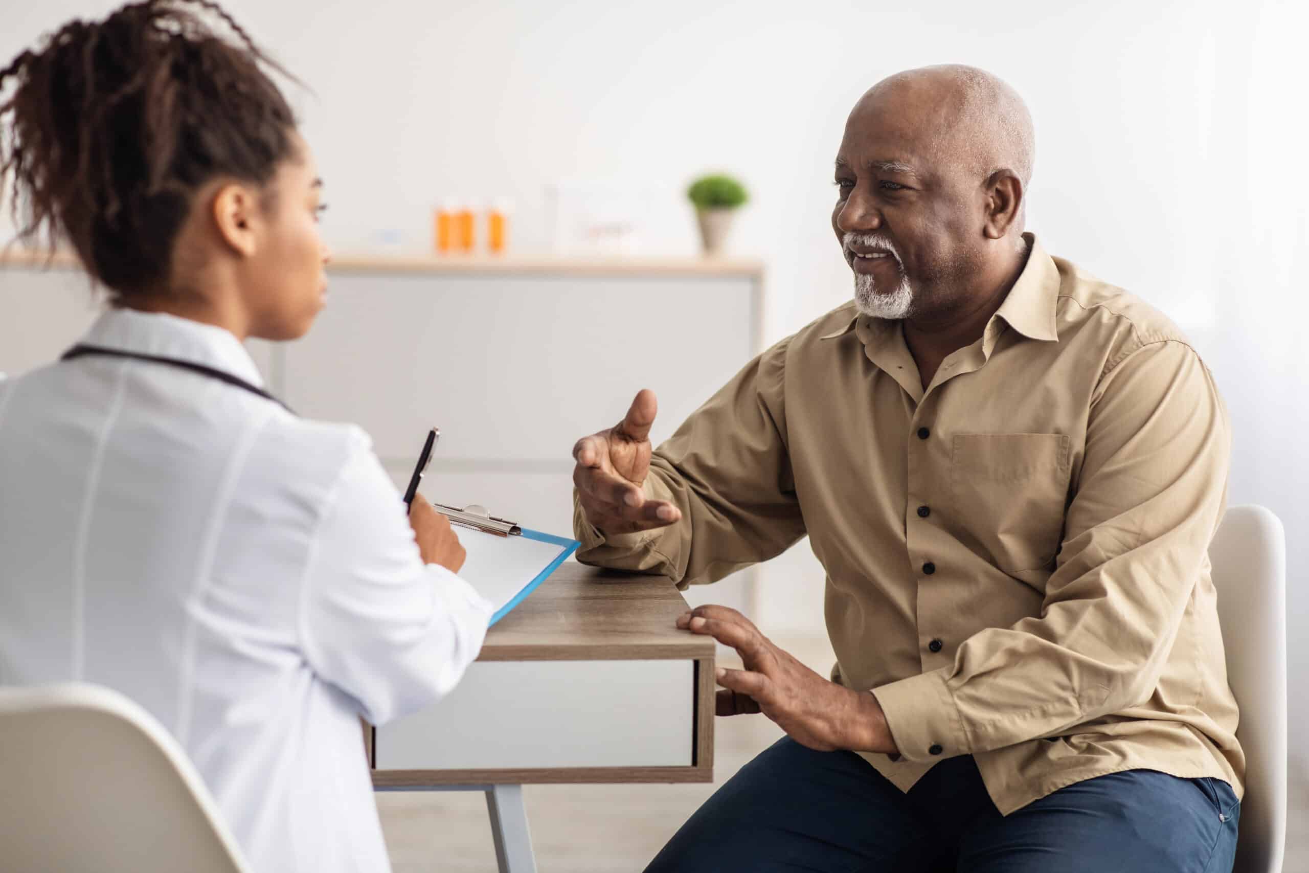 Older man sits across from a doctor at a desk, engaging in conversation as the doctor takes notes on a clipboard in a bright office setting.