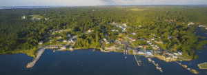 Aerial view of a small waterfront town with docks, houses, and dense green forest surrounding the shoreline.