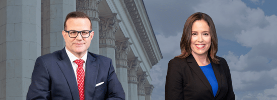 A man in a suit and tie and a woman in a blazer stand in front of a building with columns and a cloudy sky in the background.