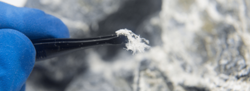 A gloved hand holds tweezers gripping fibrous white material, possibly asbestos, with rocks in the background.