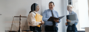 Three business professionals stand in an office, holding folders and having a discussion. A balance scale is visible in the foreground.