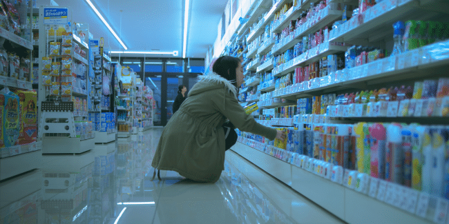 A person in a coat is crouching and selecting items from shelves in a brightly lit convenience store. Another person is visible near the entrance in the background.