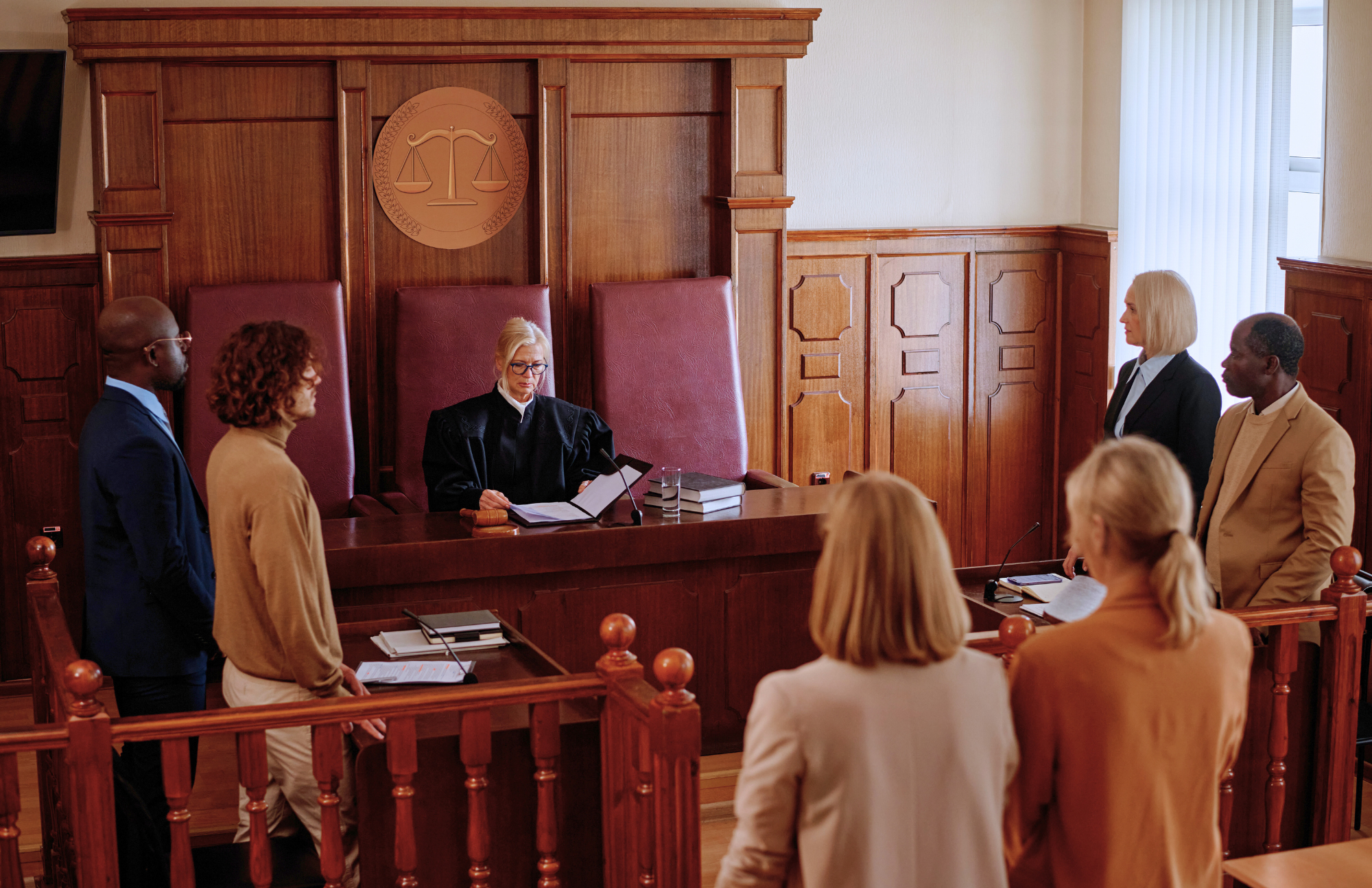 A judge sits at the bench in a courtroom while six people, three on each side, stand facing the judge. Papers and books are on the judge’s desk.