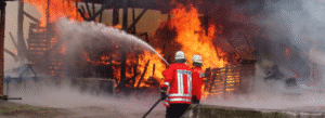 Two firefighters in red jackets spray water on a large fire consuming a wooden structure, with heavy smoke and flames visible.