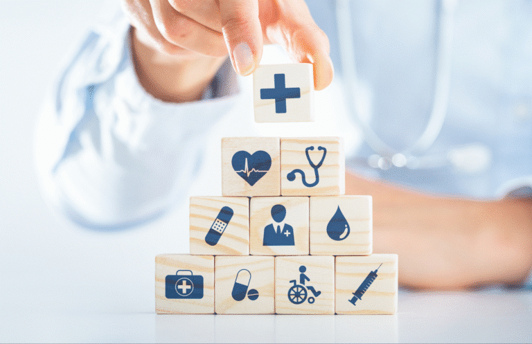 A hand places a wooden block with a medical cross symbol atop a pyramid of blocks featuring various healthcare icons, such as a heart, stethoscope, and syringe.