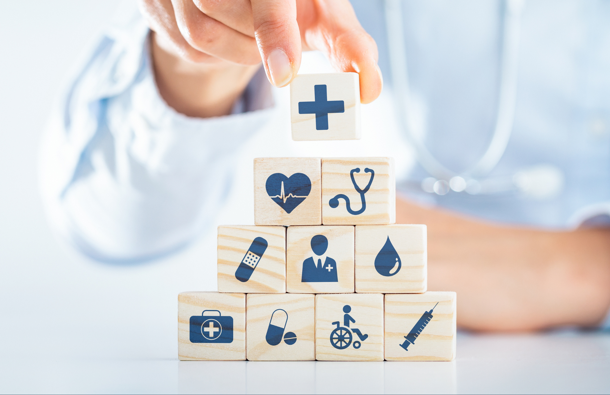 A hand places a wooden block with a medical cross symbol atop a pyramid of blocks featuring various healthcare icons, such as a heart, stethoscope, and syringe.