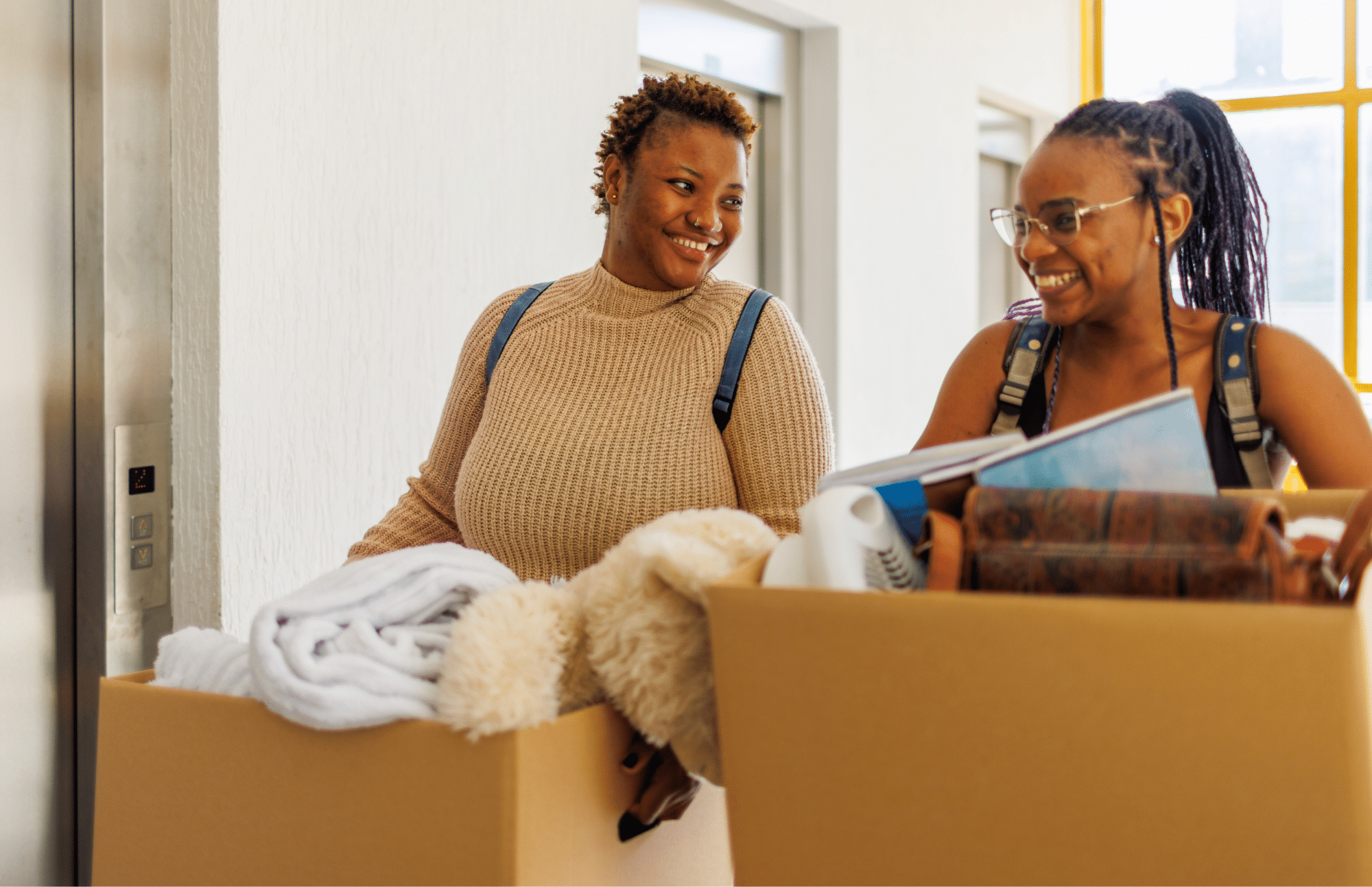 Two women carrying large cardboard boxes filled with household items smile at each other in a hallway near an elevator.