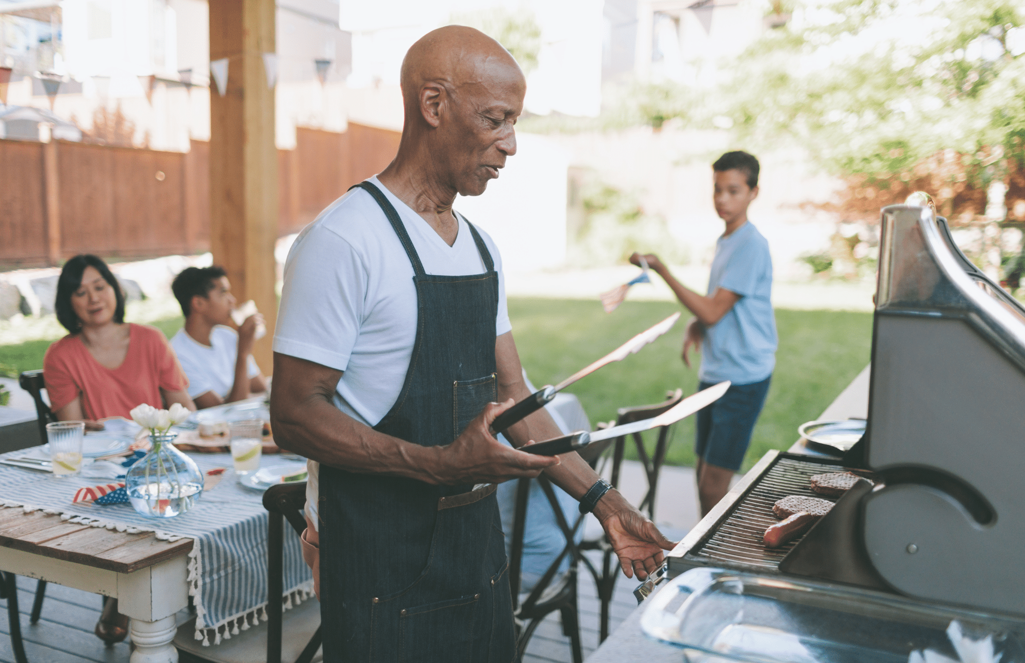 A man in an apron grills food on a barbecue in a backyard while three people sit at a table and another person stands nearby holding a plate.