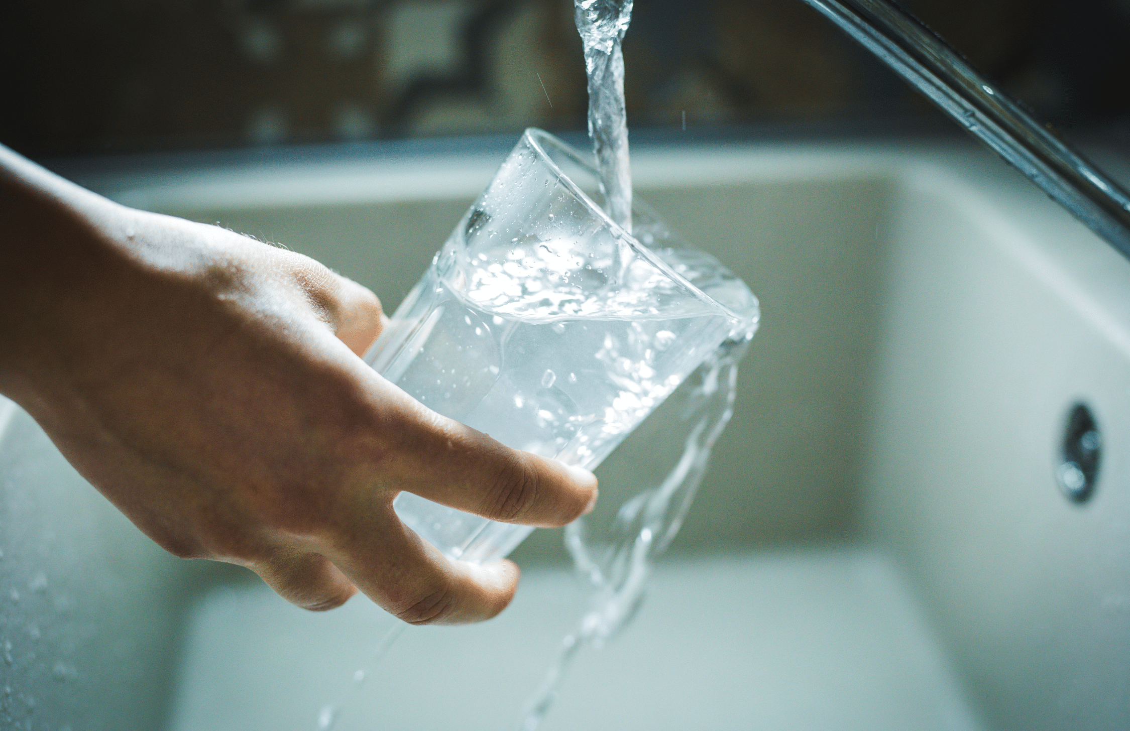 A hand holds a clear glass under a running faucet, filling it with water in a sink.