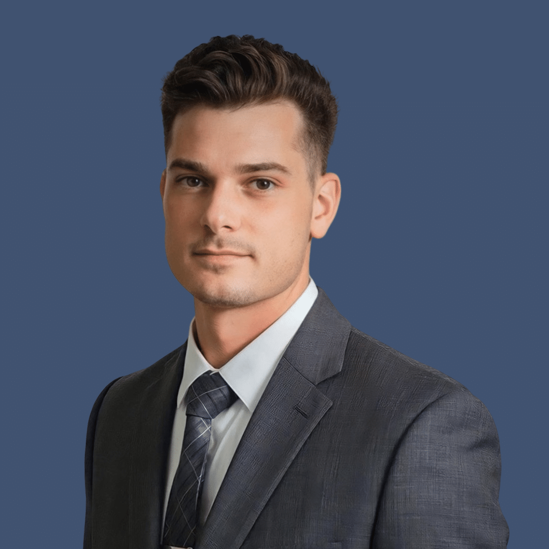 A young man in a dark suit, white shirt, and striped tie poses against a plain blue background.