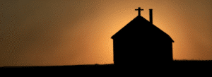 Silhouette of a small church with a cross on the roof at sunset, set against a gradient orange and brown sky.